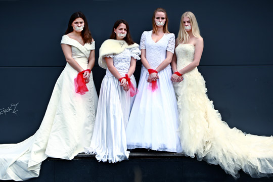 4 unge Frauen in weißen Hochzeitskleidern mit langem Schleier, die Münder der Mädchen sind mit Pflaster zugeklebt, die Arme mit rotem Stoff zusammengebunden
