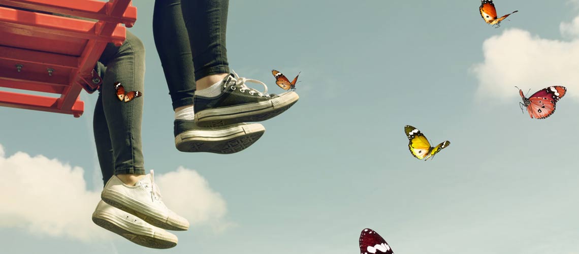 Die Beine und Füße zweier Mädchen, die Turnschuhe anhaben, sind zu sehen, im Hintergrund der blaue Himmel, im Vordergrund umherfliegende Schmetterlinge